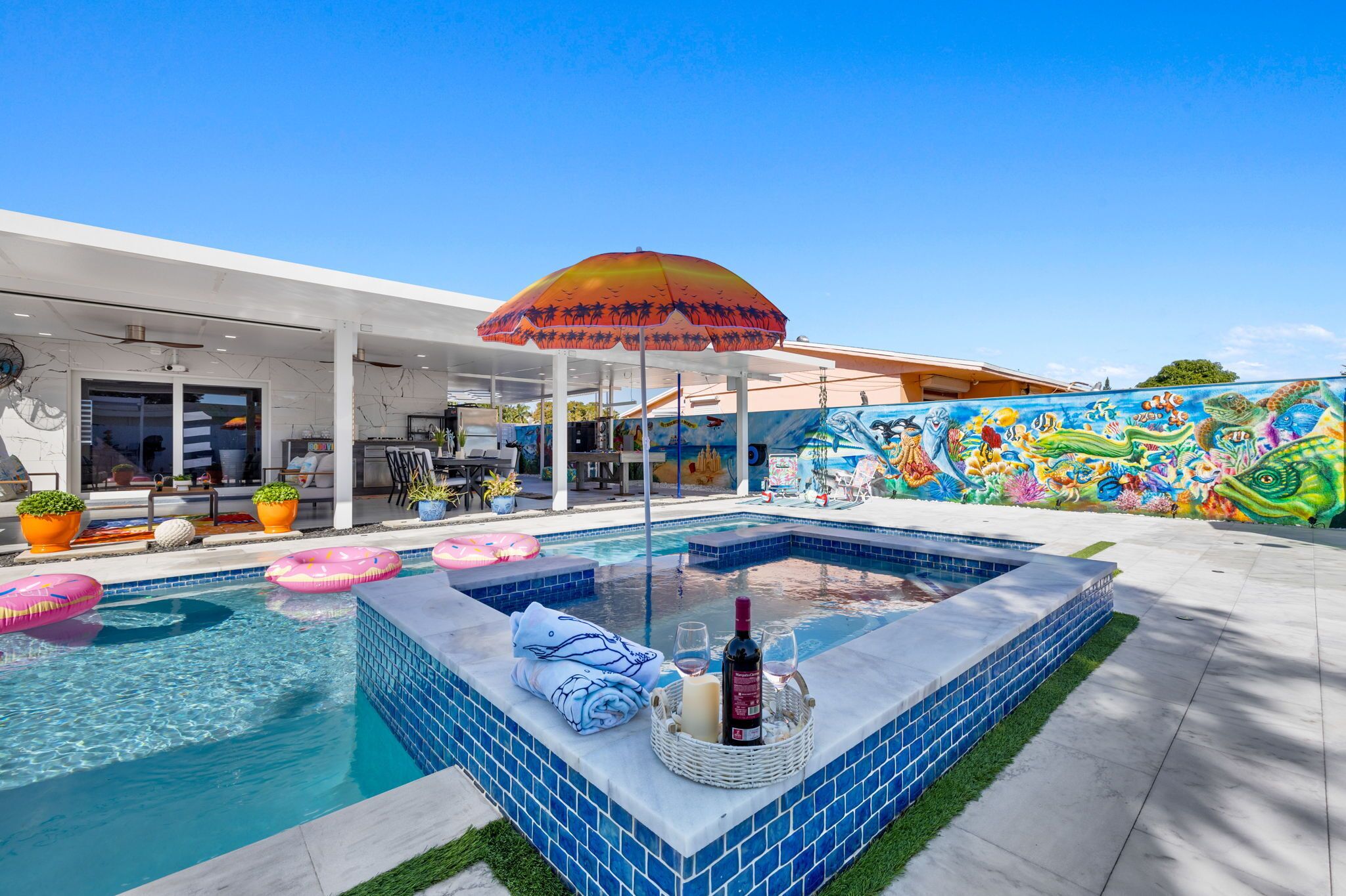 Une piscine avec un jacuzzi, entourée d’un patio couvert doté de meubles d’extérieur et d’un mur décoré d’une fresque.