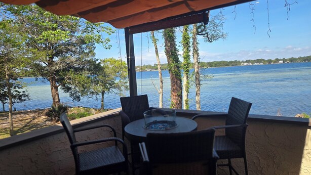 Een overdekt terras met een tafel en stoelen, met uitzicht op een waterpartij met bomen en gebouwen in de verte.