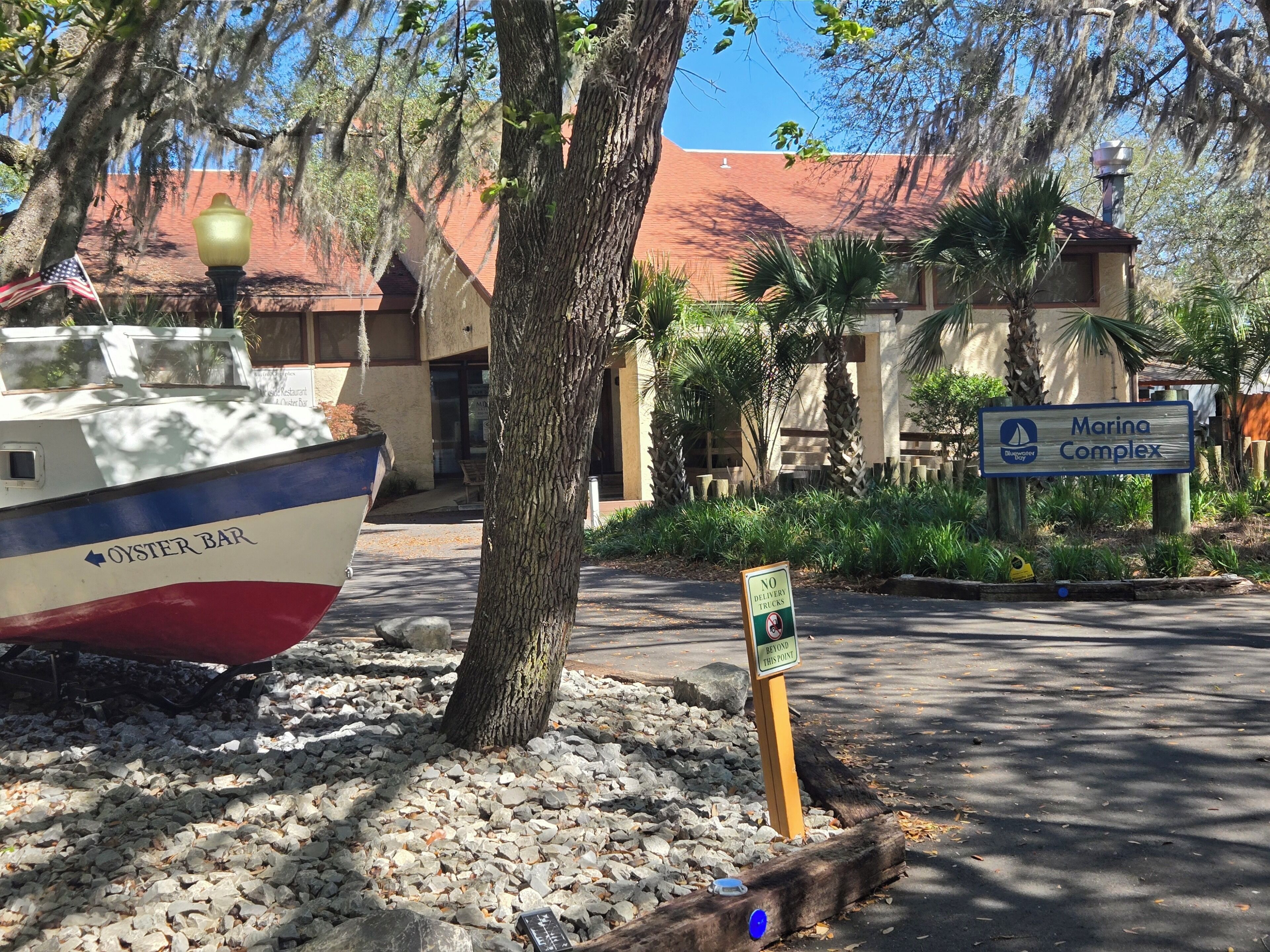 A boat labeled 'Oyster Bar' parked near a tree, a sign for 'Marina Complex,' and a 'NO DELIVERY TRUCKS' sign.