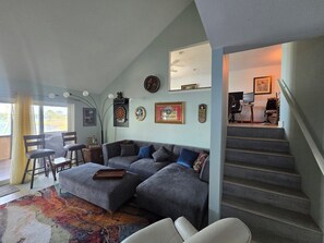 Ein Wohnzimmer mit einem Ecksofa, einer Dartscheibe und einem Fernseher. Eine Treppe führt zu einer weiteren Ebene.