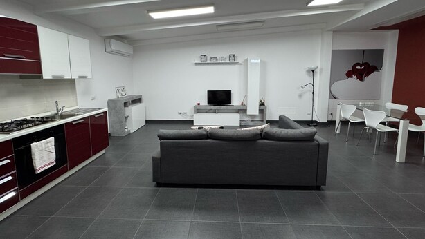 A modern kitchen with a gas stove, a dining area with a table and chairs, a living room with a sofa, and a TV. - Vertigo Casoria (Casoria)