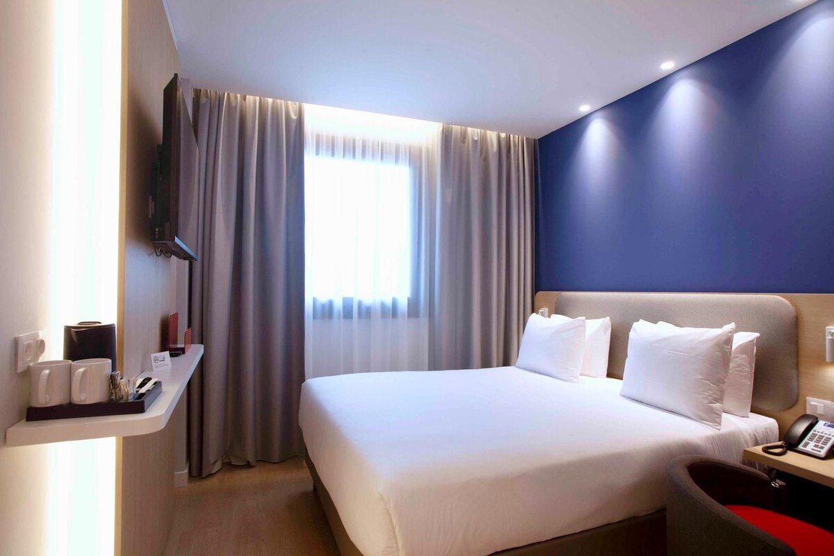 Une chambre d’hôtel avec un grand lit, une table de chevet avec un téléphone, un téléviseur fixé au mur et une fenêtre avec des rideaux.