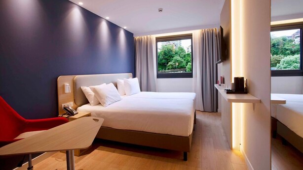 Habitación de hotel moderna con cama, escritorio, silla, televisor y ventana con vista a follaje.