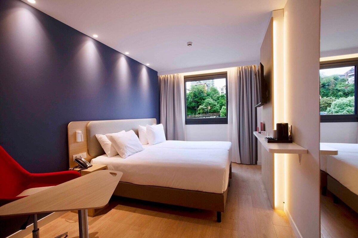 Une chambre d’hôtel moderne équipée d’un lit, d’un bureau, d’une chaise, d’une télévision et d’une fenêtre donnant sur un paysage verdoyant.