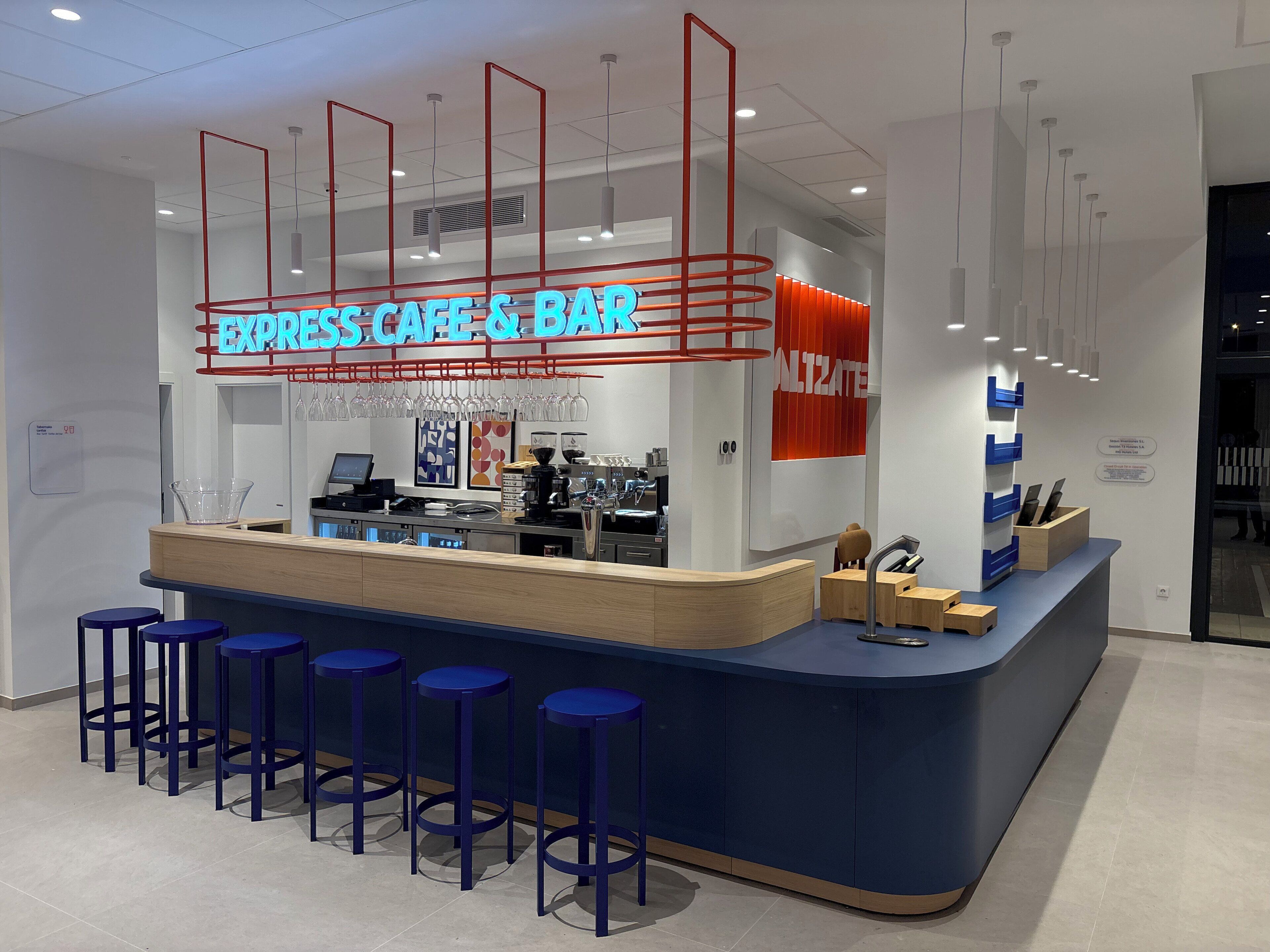 Un moderno café y bar con un letrero de neón, taburetes, una máquina de café y una barra con un ordenador.