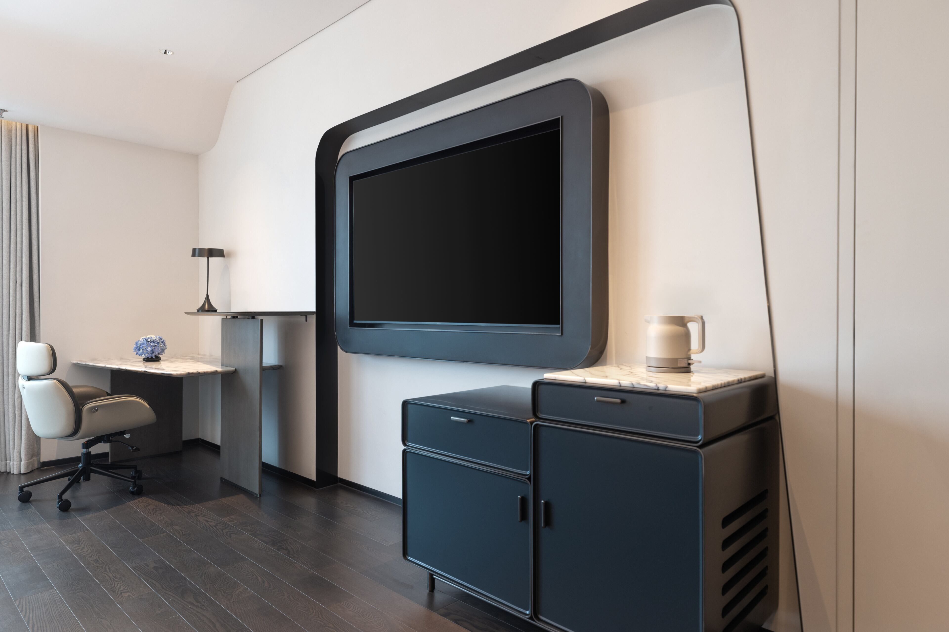 A modern hotel room with a large flat-screen TV, a desk with a lamp, a chair, and a cabinet.