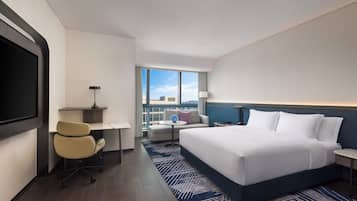 A hotel room with a large bed, a desk, a chair, a TV, a lamp, and a view of the city.