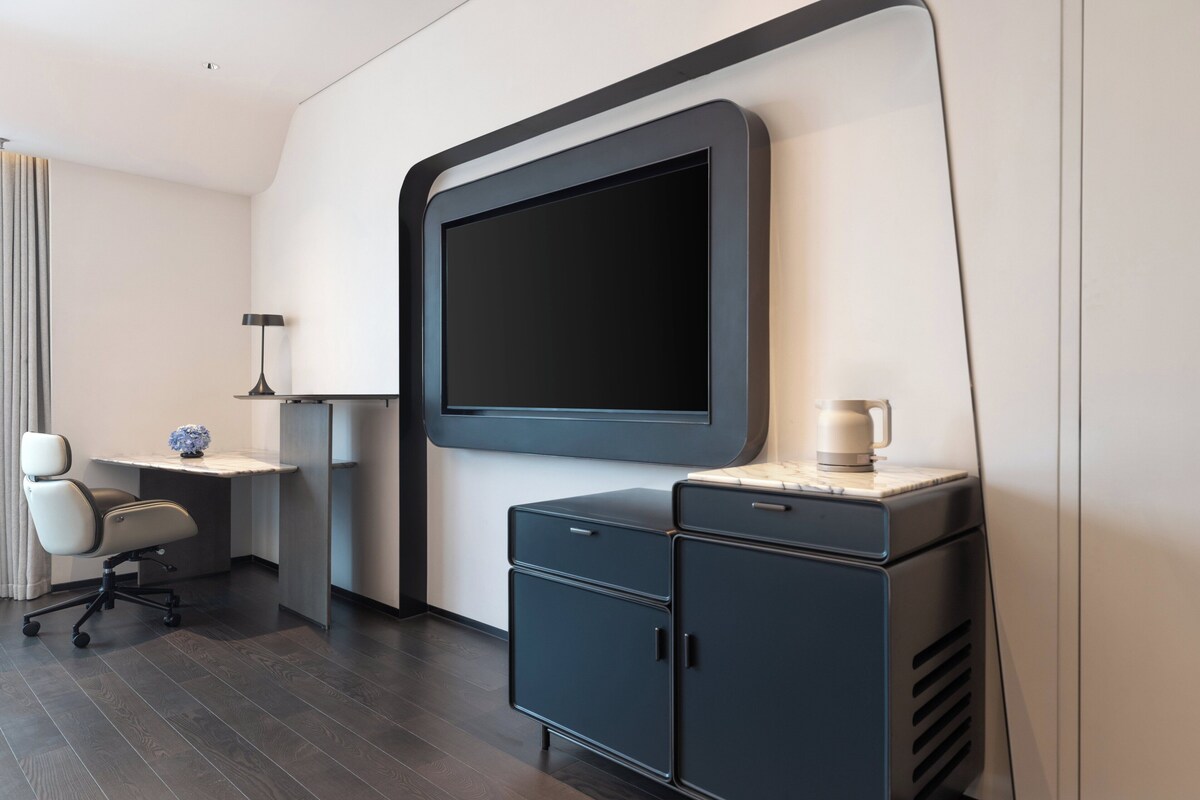A modern hotel room with a large flat-screen TV mounted on the wall, a desk with a lamp, a chair, and a small cabinet.