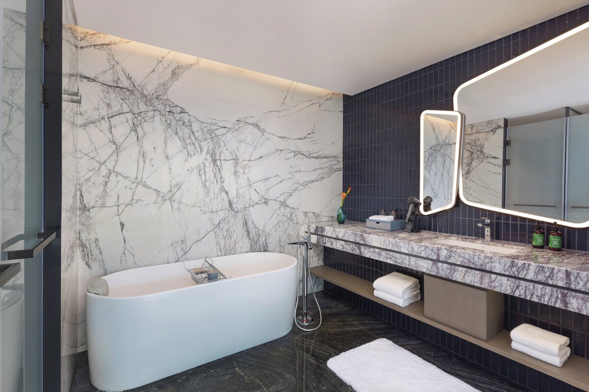 A modern bathroom with a freestanding bathtub, a large mirror with integrated lighting, a marble wall, and a dark tiled floor.