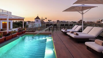 Una piscina en la azotea con sillones de descanso y vistas a la ciudad al atardecer.