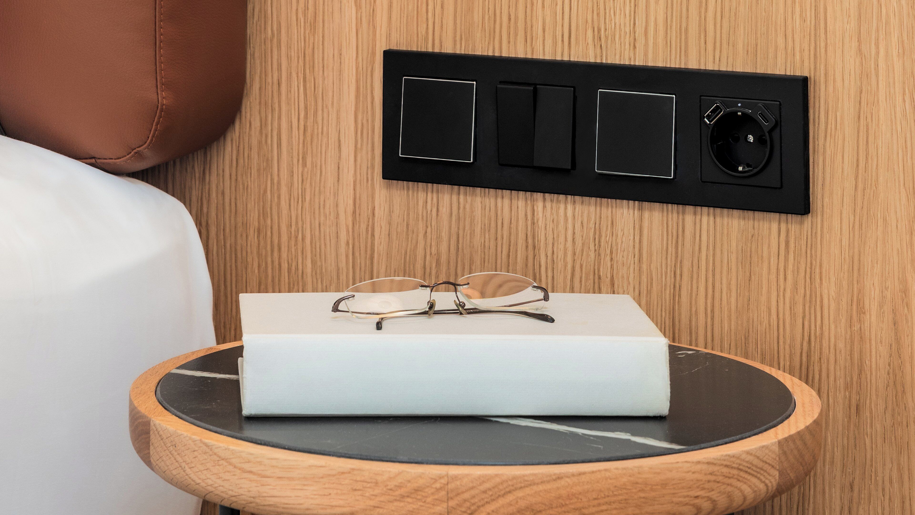 A bedside table with a pair of glasses and a book, a power outlet, and a switchboard on a wooden wall.