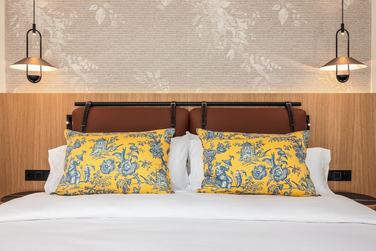 Un lit avec du linge de lit blanc, deux oreillers jaunes à motifs bleus et une tête de lit en bois.