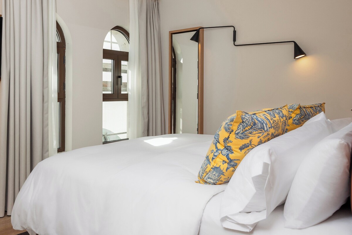 A hotel room with a bed, white bedding, a flat-screen TV mounted on the wall, a mirror, and a bedside lamp.