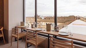 A dining area with a table set for two, a view of a cityscape through large windows, and a wooden chair with a woven seat. - Casa Del Compositor, Vignette Collection by IHG (Jerez de la Frontera)