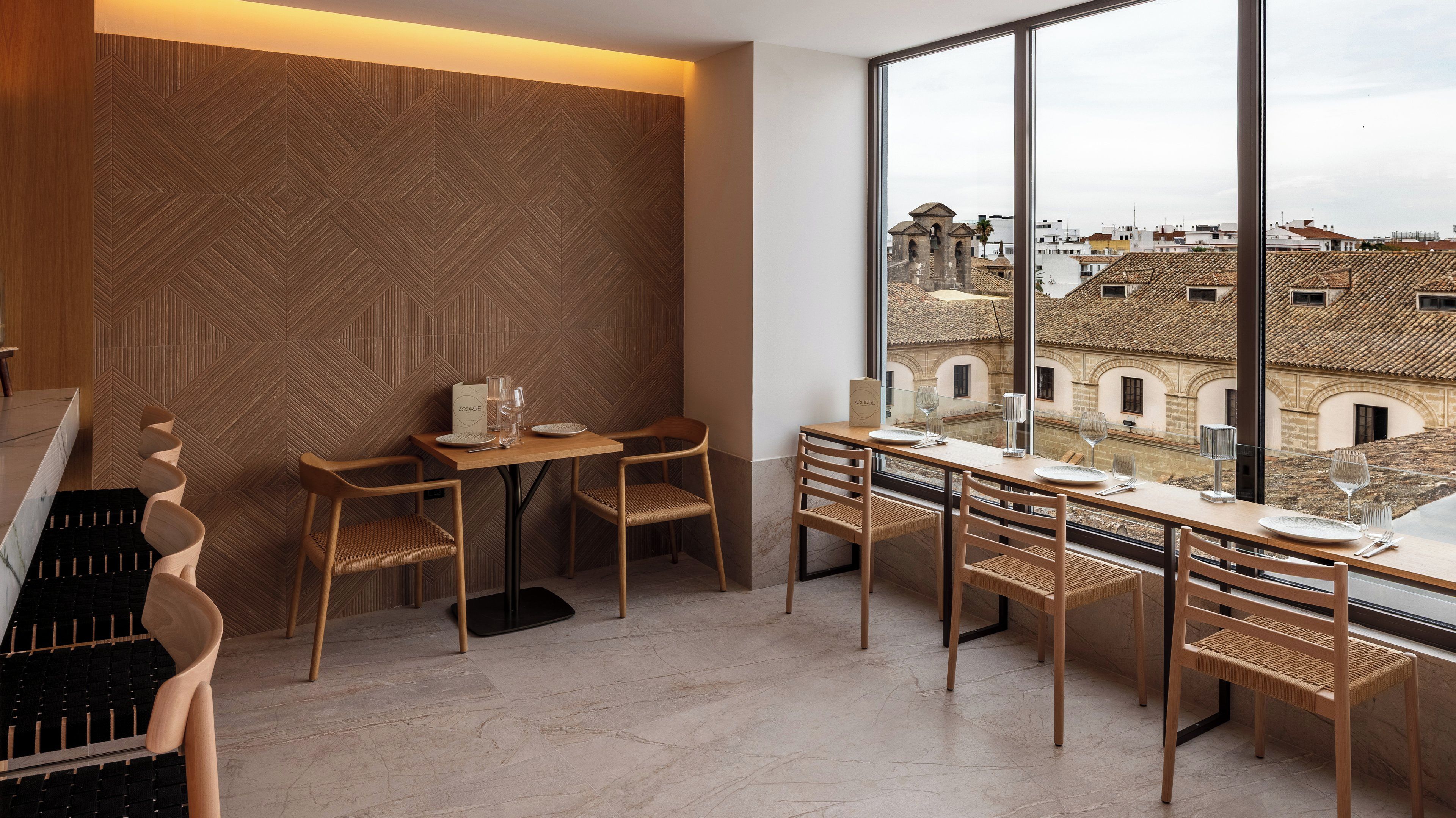 Un coin repas avec une table dressée pour deux personnes, des chaises en bois et une vue sur les bâtiments à travers de grandes fenêtres.