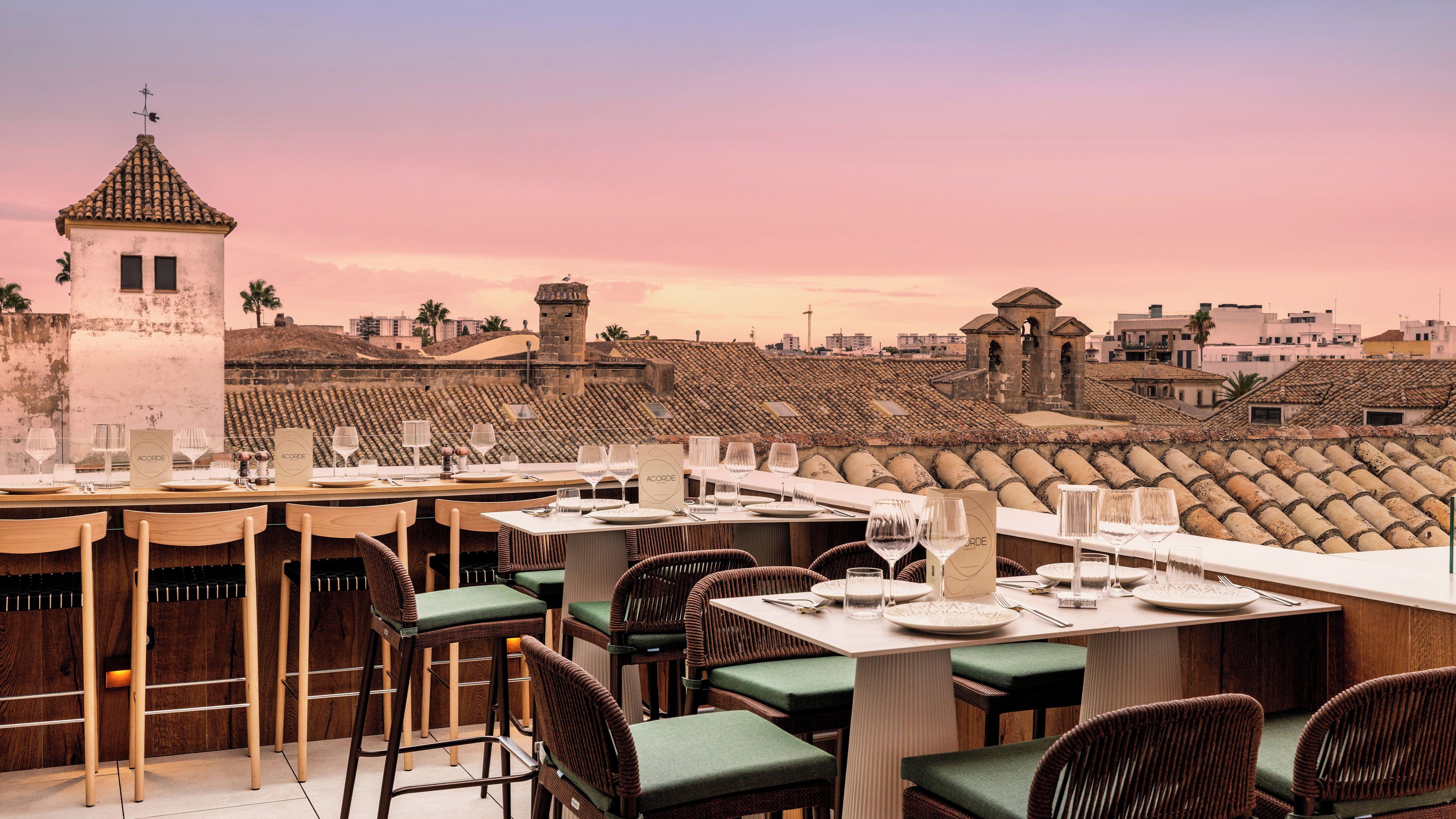 Een dakrestaurant met gedekte tafels, met uitzicht op een historisch stadsbeeld met terracotta daken en een toren.