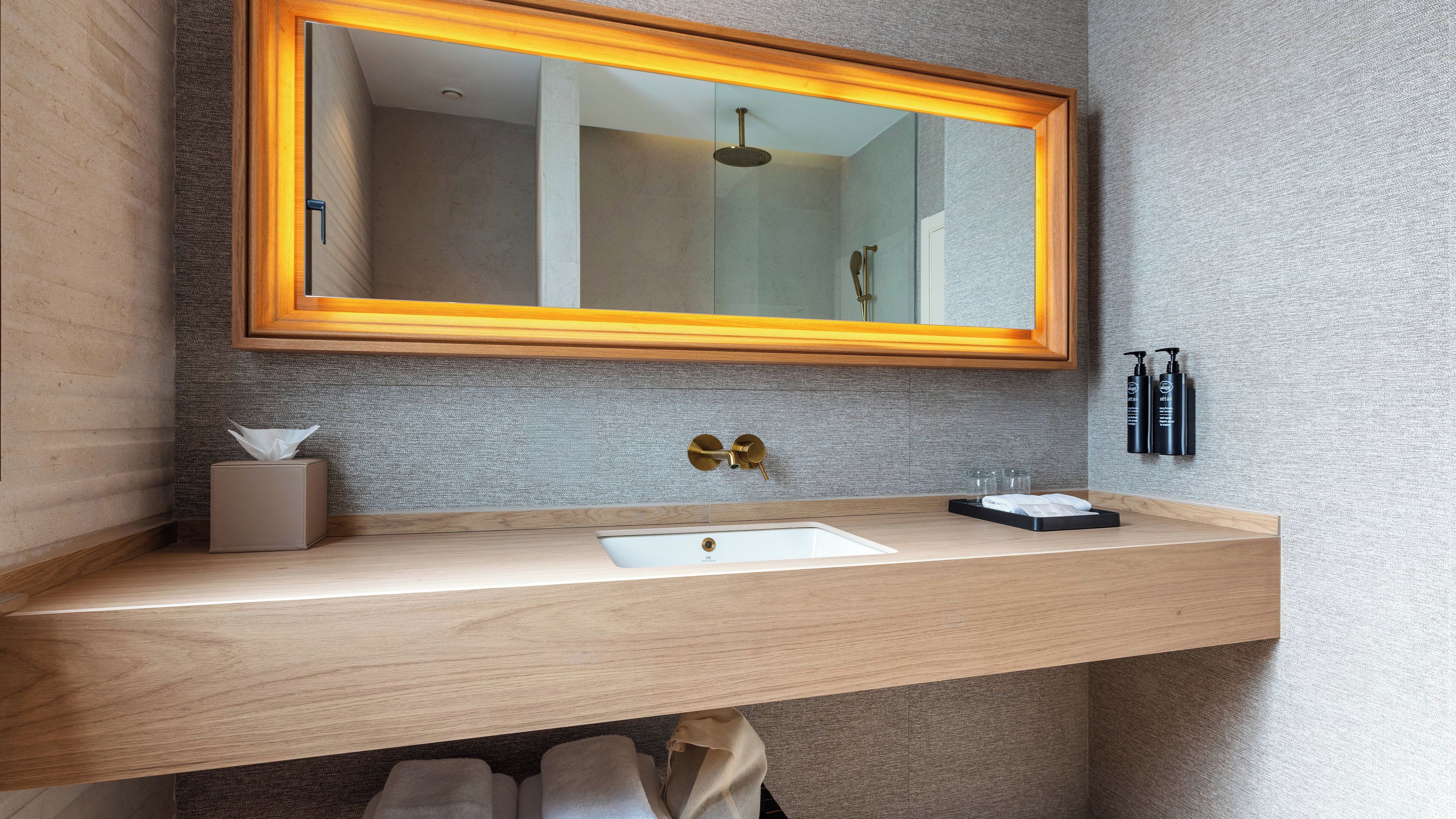 A bathroom with a wooden vanity, a mirror with a golden frame, a towel rack, and a bottle of shampoo.
