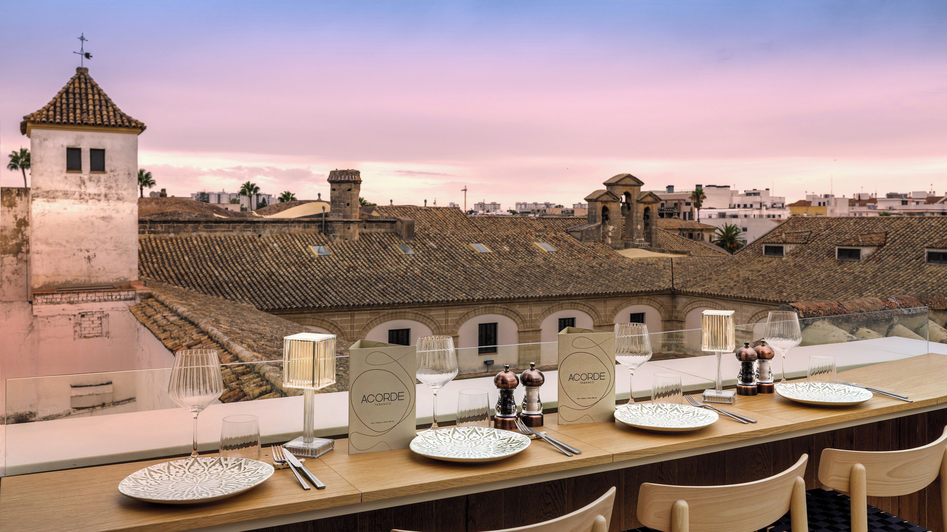 Una mesa de comedor dispuesta con platos, vasos y cubiertos, con vistas a un edificio histórico de tejados de azulejos.