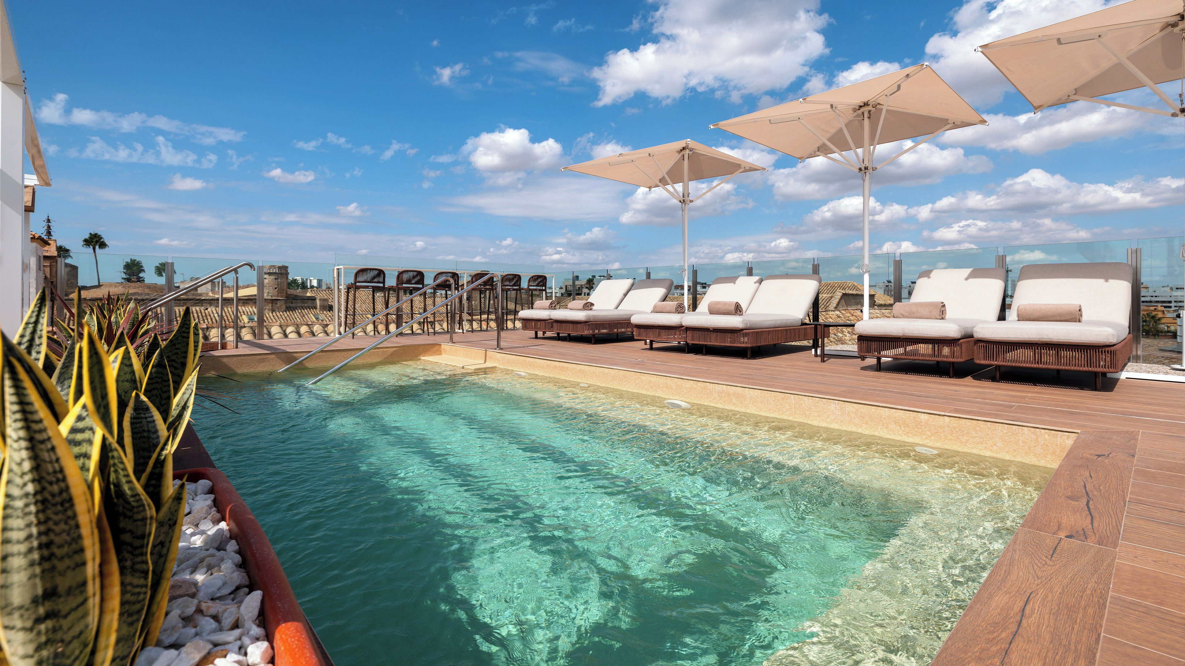Un área junto a la piscina con sillones de descanso y sombrillas, agua cristalina y vistas del paisaje urbano.