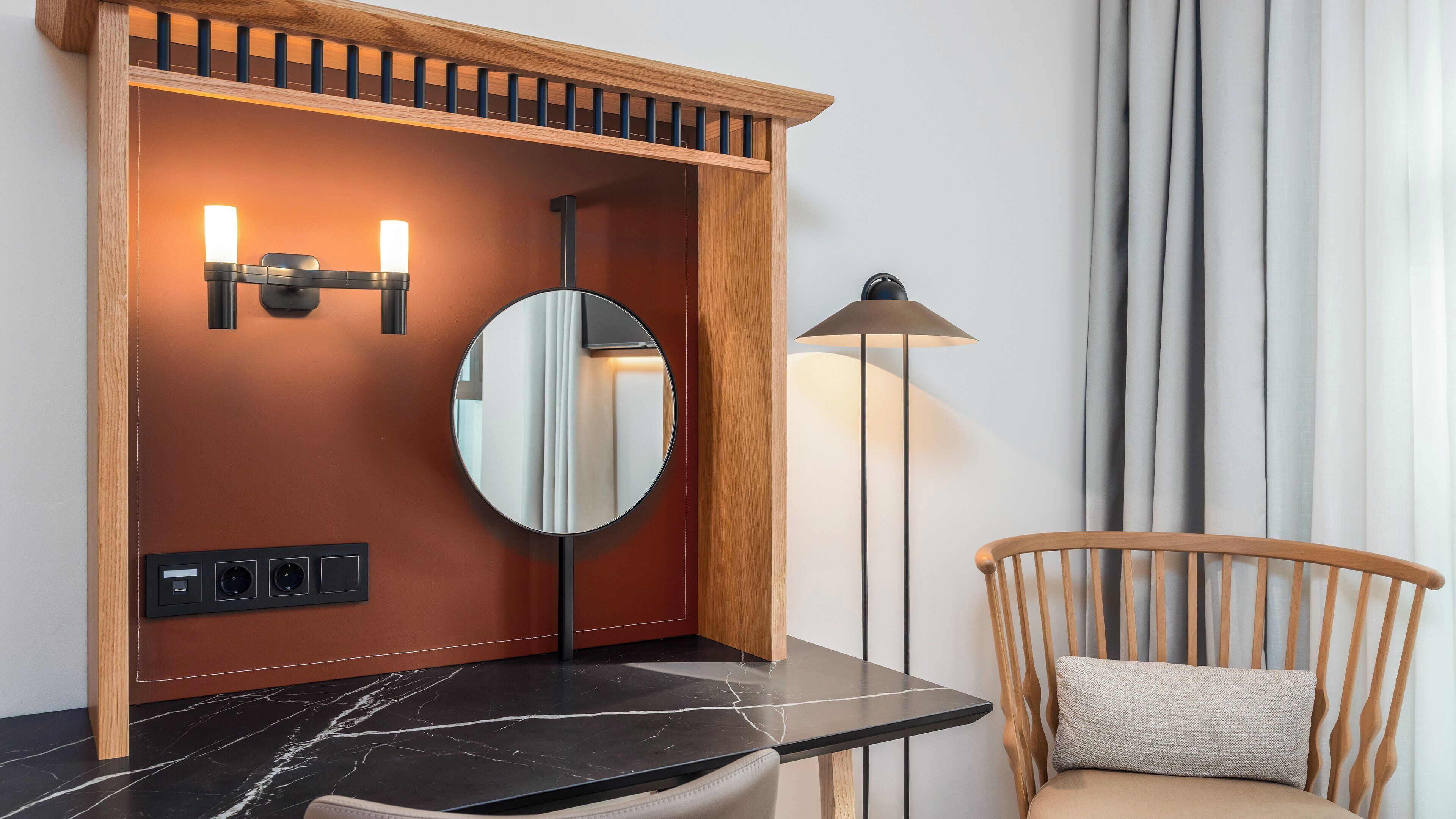 A bathroom with a wooden vanity, a round mirror, two wall-mounted lights, a floor lamp, and a chair with a cushion.