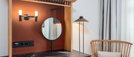 A bathroom with a wooden vanity, a round mirror, two wall-mounted lights, a floor lamp, and a chair with a cushion.