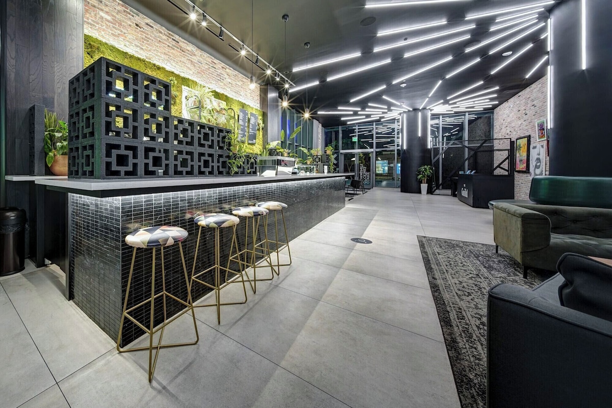 Zona de bar moderna con salpicadero de azulejos blanco y negro, una pared verde con plantas y un área de asiento con taburetes de bar.