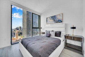A modern bedroom with a large bed, a nightstand, a wall-mounted lamp, and a city view through a large window. - Alea Miami (Miami)