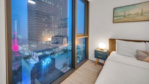 Ein modernes Hotelzimmer mit großem Fenster und Blick auf die Stadt, einem ordentlich bezogenen Bett und einem kleinen Nachttisch mit Lampe.