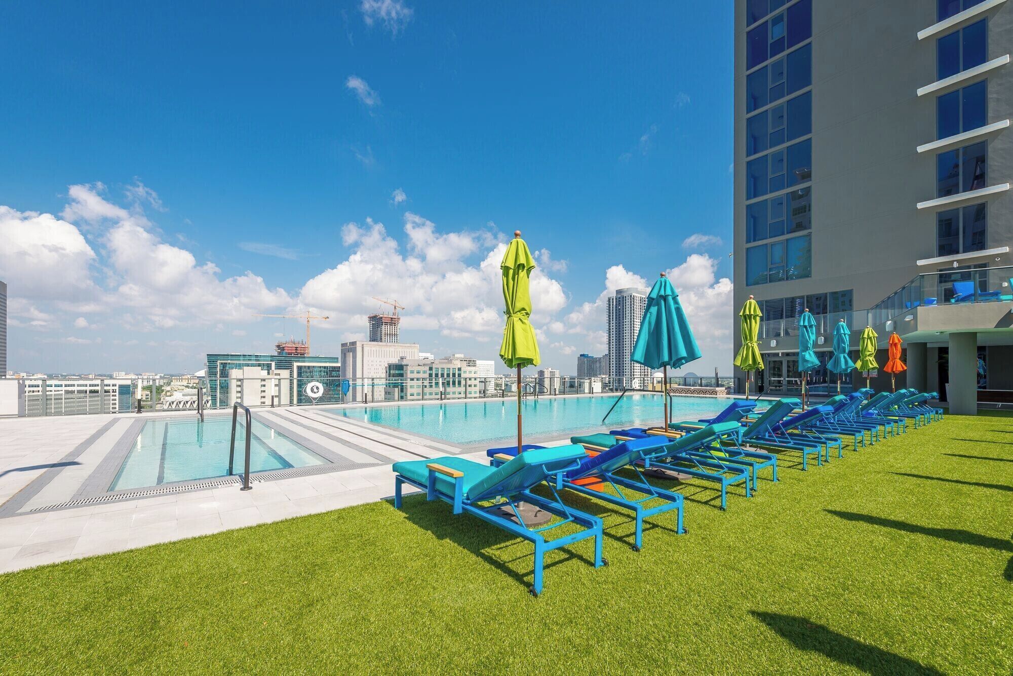 A rooftop pool area with blue lounge chairs and green umbrellas, surrounded by a grassy lawn and modern buildings in the background.