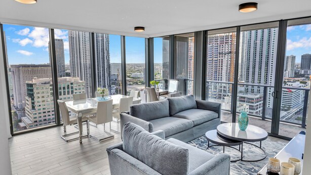 Ein modernes Wohnzimmer mit einem grauen Sofa, einem runden Couchtisch und einem Essbereich mit weißem Tisch und Stühlen. Große Fenster bieten Blick auf die Stadt.