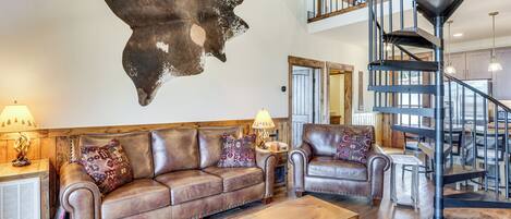 A spacious living area with a brown leather sofa, a wooden coffee table, and a large wall-mounted animal hide. A spiral staircase with a metal railing is visible in the background.