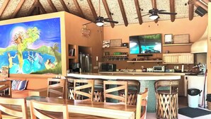 Una habitación con una mesa y sillas de madera, un área de cocina con electrodomésticos, un televisor y un mural con una escena tropical.