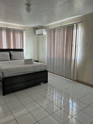 A bedroom with a bed, curtains, an air conditioning unit, and a tiled floor.