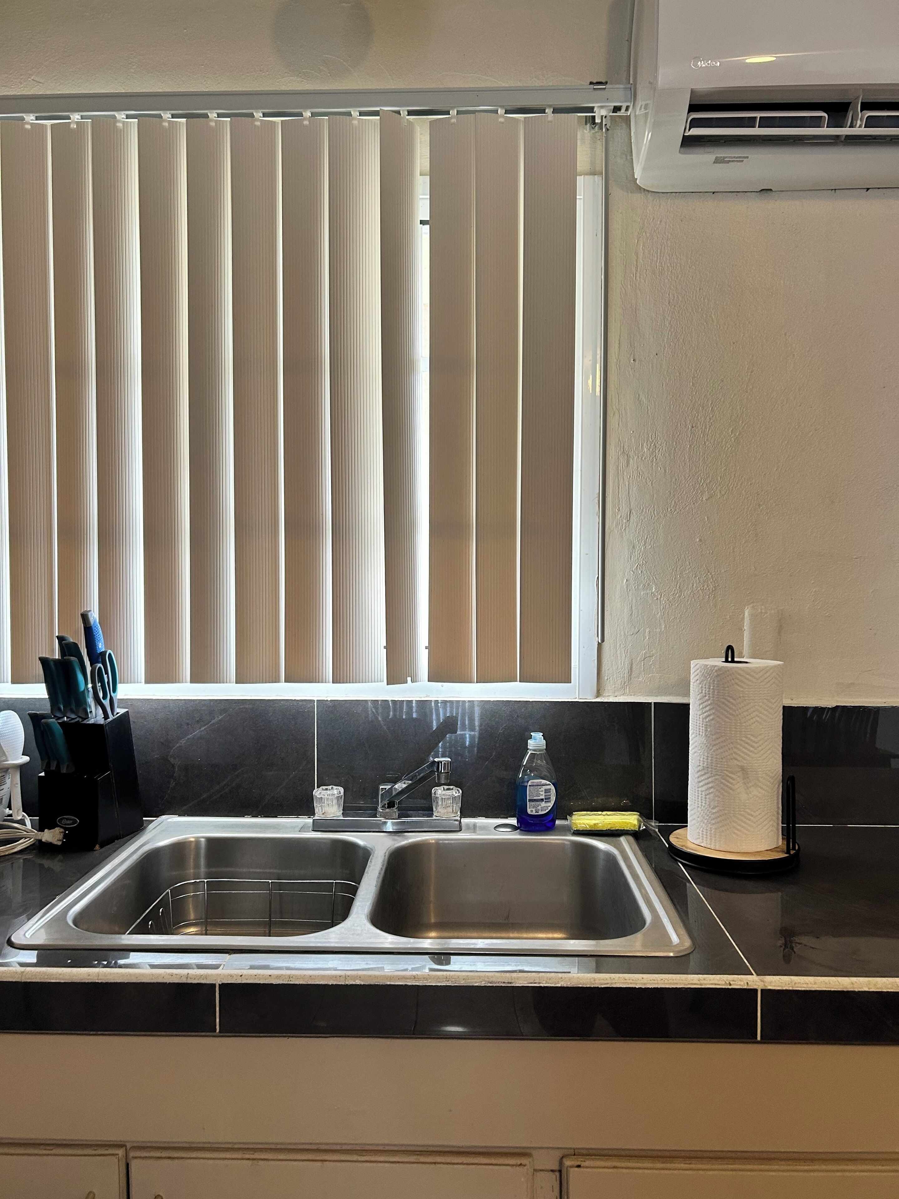 A kitchen with a double sink, a window with vertical blinds, a paper towel holder, and a dish soap bottle.