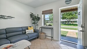 Ein Wohnzimmer mit grauem Sofa, einem Couchtisch mit einer Zeitschrift, einer Topfpflanze und einem Fenster mit Blick auf eine grüne Rasenfläche und ein Gebäude.