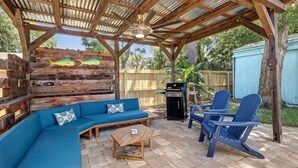 Eine überdachte Terrasse im Freien mit Holzboden, einem blauen Sofa, zwei Adirondack-Stühlen und einem kleinen Tisch.