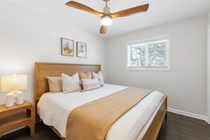 Une chambre avec un lit en bois, une table de chevet avec une lampe, un ventilateur de plafond et deux tableaux encadrés accrochés au mur.
