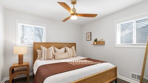 Un dormitorio con una cama de madera, un ventilador de techo, una lámpara de noche, una ventana con persianas y un pequeño estante con objetos decorativos.
