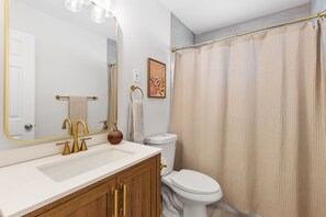 Ein Badezimmer mit Waschbecken in Weiß, goldenen Armaturen, einem Holzkästen, einer weißen Toilette und einem beige farbenen Duschvorhang.