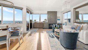A spacious living room with a large window overlooking the sea, a dining area with a wooden table and chairs, and a cozy seating area with a blue sofa and a fireplace. - Ocean View Home | Walk to Beach & Downtown MB (Manhattan Beach)