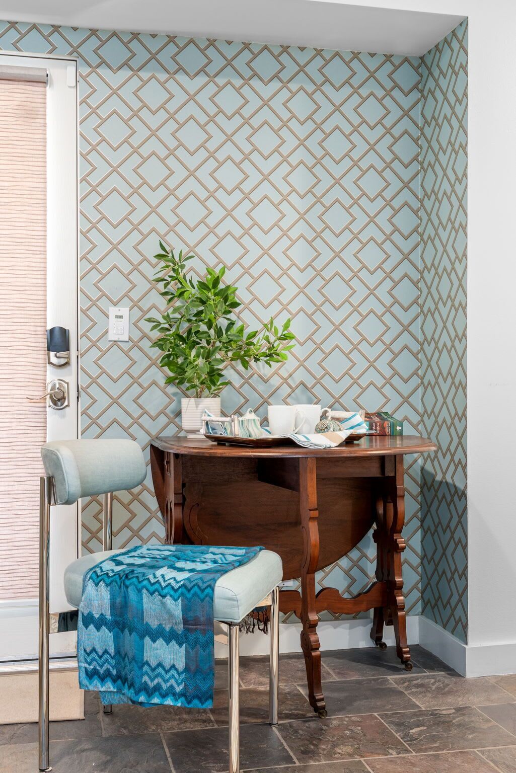 A corner room with a patterned wallpaper, a wooden table with a plant, a chair, and a blue throw blanket.
