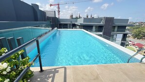 Una piscina en la azotea con barandilla de vidrio, vista panorámica de la ciudad y una grúa de construcción al fondo.