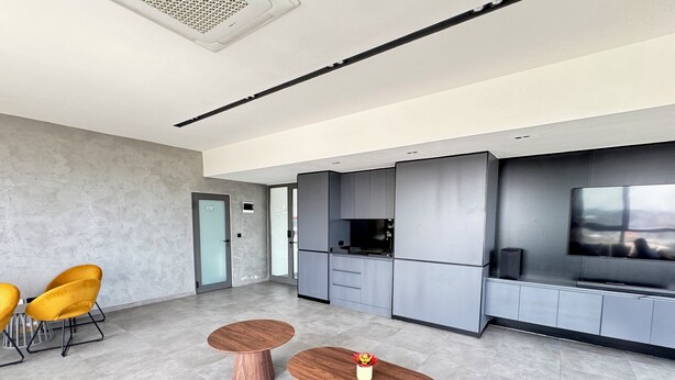 Una cocina moderna con gabinetes grises, horno empotrado y televisor de pantalla plana. En la sala de estar, hay dos mesas de madera con sillas.
