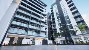 Un edificio moderno de varios pisos con una entrada destacada, ventanas amplias y un área ajardinada con palmeras.