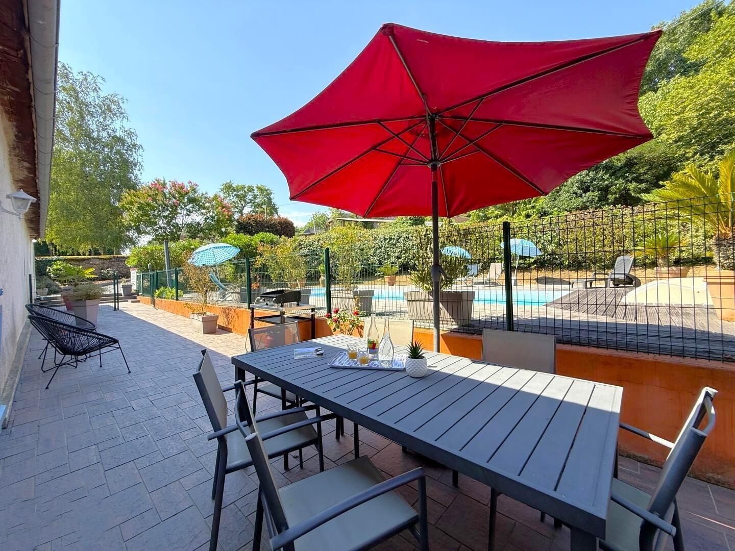 Un patio con mesa y sillas, un paraguas rojo y una piscina al fondo.