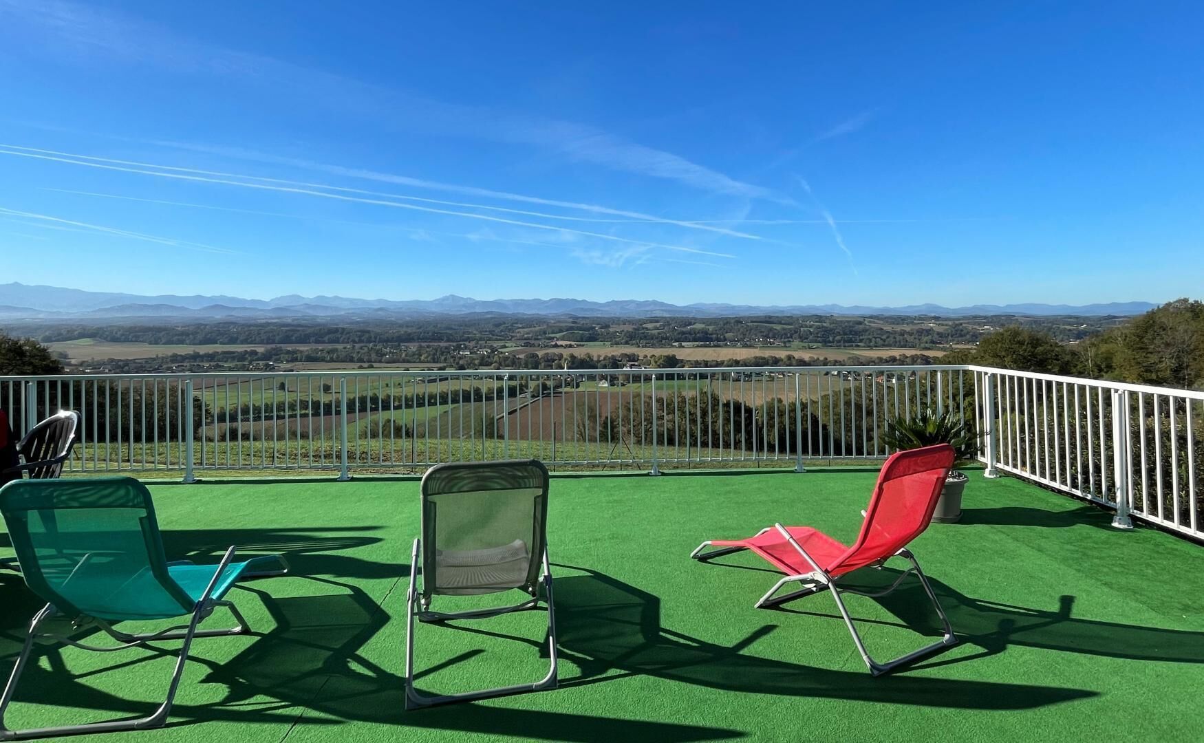 Une terrasse dotée d’un revêtement de sol en gazon synthétique vert, de chaises en métal et d’une rambarde blanche, offrant une vue imprenable sur un paysage pittoresque.