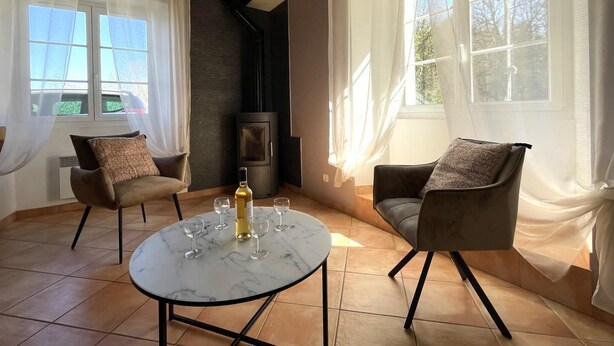Ein gemütlicher Raum mit zwei Sesseln, einem runden Tisch mit Weingläsern und einer Flasche. Große Fenster mit durchsichtigen Vorhängen, ein Kamin und ein Fliesenboden sind ebenfalls vorhanden.
