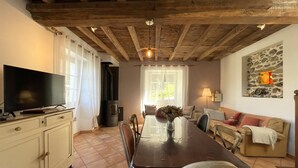 Ein gemütliches Wohnzimmer mit Holzbalkendecke, einem Fernseher auf einem Schrank, einem Esstisch mit Stühlen, einer Couch und einer Steinwand mit einer Kerze.