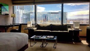 Ein Hotelzimmer mit großem Fenster und Blick auf die Stadt, einem Bett, einem Sofa, einem Couchtisch und einem Schreibtisch mit Lampe.