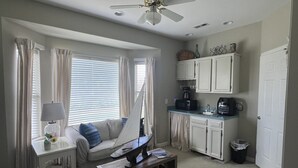 Ein gemütlicher Wohnbereich mit Küchenzeile, Deckenventilator, einer Couch mit Kissen, einem kleinen Tisch und einem Fenster mit Vorhängen.